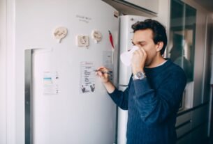 Man drinks coffee while writing a to-do list on a kitchen refrigerator.