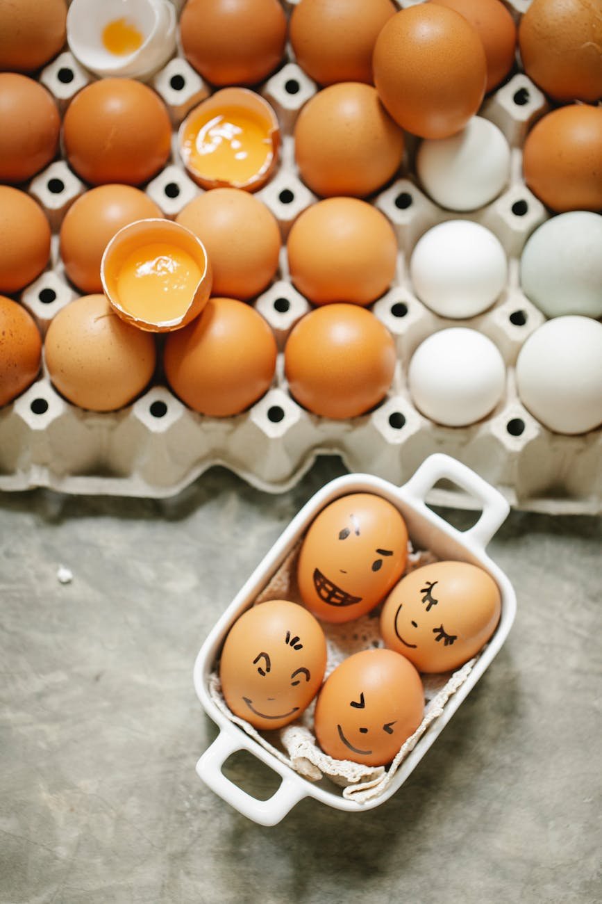 From above of square bowl with brown chicken eggs with painted faces placed near carton box with assorted production in kitchen